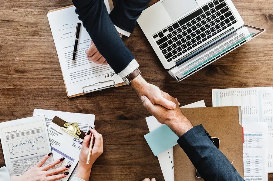 Aperto de mãos sobre uma mesa com contrato, notebook e relatórios financeiros